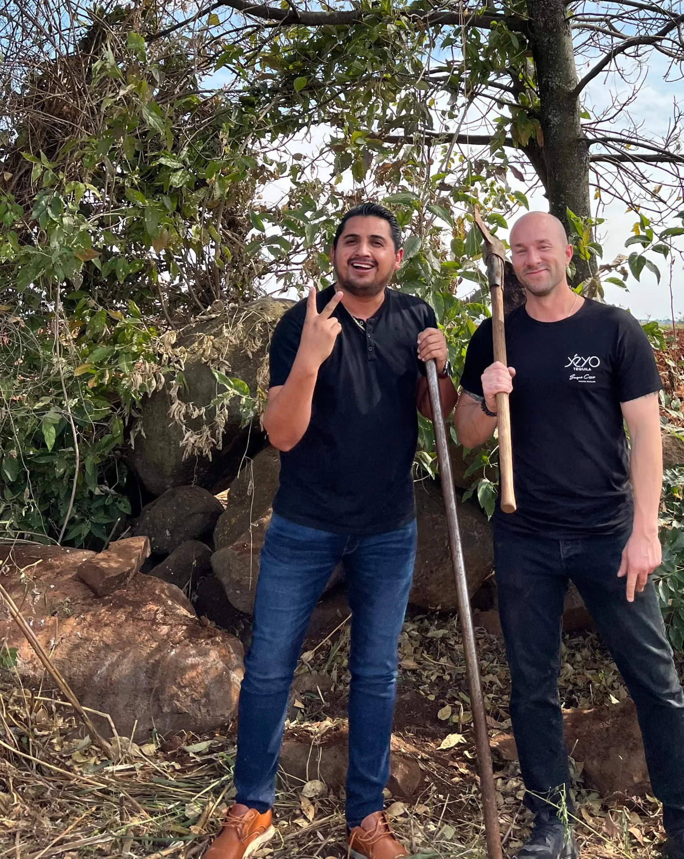 Master Distiller Sergio Cruz and founder Jon Bullinger standing with digging tools beneath a tree in Arandas, Jalisco