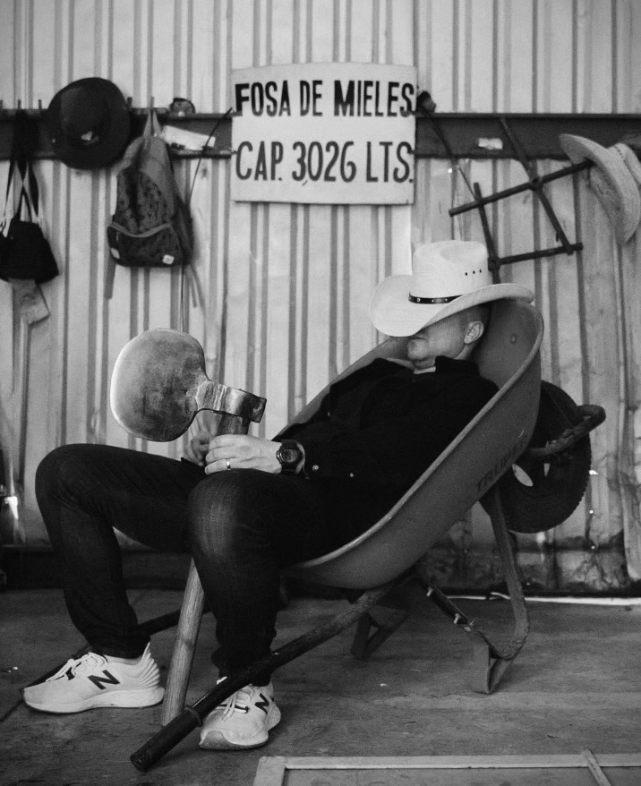 Black-and-white photo of a man in a cowboy hat resting in a wheelbarrow beneath a Fosa de Mieles sign